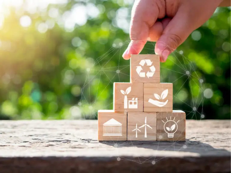 Mão colocando um bloco de madeira com um símbolo de reciclagem no topo de uma pilha de outros blocos com ícones relacionados a ESG, como fábrica, folha, prédio, turbina eólica e lâmpada, com um fundo verde borrado